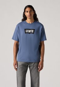Mėlynas medvilnės marškinėlis su juodu Levi's logotipu ant kreivos emblemos. Trumpi rankovės, apvalus kaklas ir laisvas prigludimas, derinami su pilkais džinsais.