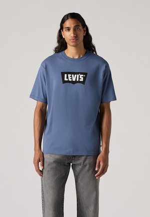 Mėlynas medvilnės marškinėlis su juodu Levi's logotipu ant kreivos emblemos. Trumpi rankovės, apvalus kaklas ir laisvas prigludimas, derinami su pilkais džinsais.