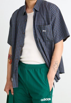 Persoon draagt een blauwbruin geruit kort mouwen overhemd over een wit hemd en groene Adidas broek, met tatoeages op beide armen.
