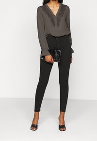 Blusa grigia scuro con dettagli in pizzo sul collo a V, abbinata a jeans skinny neri. La modella tiene una pochette nera e indossa tacchi aperti neri.