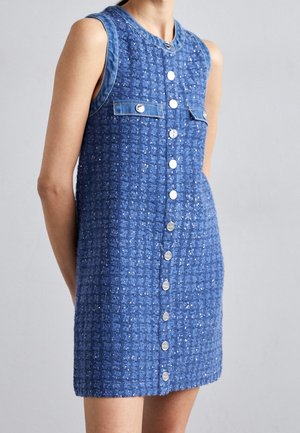 Vestido mini sin mangas azul texturizado con botones plateados en la parte delantera y dos bolsillos en el pecho con botones, llevado por una persona sobre un fondo liso.