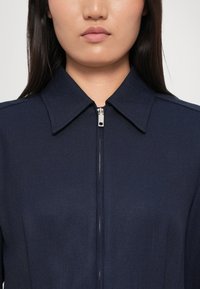 Námořnická modrá zipová bunda s límečkem a texturovanou tkaninou. Obsahuje stříbrný zip a strukturovaná ramena.