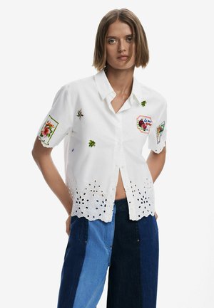 Camicia bianca a maniche corte con toppe ricamate, orlo in pizzo e design con bottoni. Presenta un colletto classico e dettagli floreali.