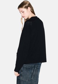 Jongere met lang haar draagt een losse zwarte gebreide trui en vervaagde blauwe jeans, kijkt omlaag en iets opzij tegen een witte achtergrond.