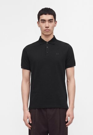 Junger Mann trägt ein schlichtes schwarzes kurzärmeliges Poloshirt mit Knöpfen und steht vor einem weißen Hintergrund.