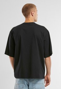 Černé bavlněné tričko s volným střihem, spadlými rameny a krátkými rukávy. Zadní strana má čistý design bez viditelného zdobení.
