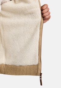 Prenda beige con cremallera y interior de forro polar crema y suave, con dobladillo de canalé en color marrón claro. Presenta una cremallera de bronce suave y una pequeña pestaña de cuero.