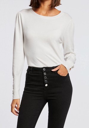 Kobieta ubrana w biały sweter z długim rękawem z zapięciem na guziki na mankietach oraz czarne, wysokowalne dżinsy z pięcioma guzikami z przodu, z ręką w kieszeni.