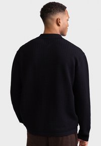 Camisola preta texturada com padrão em zig-zag e gola, com mangas compridas e ajuste descontraído. Mostrada de costas.