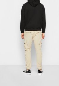 Svart hoodie, beige cargobyxor med sidofickor och svarta sneakers. Tyget verkar mjukt, med elastiska muddar och midjedetaljer.