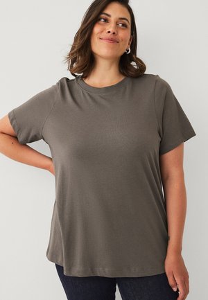 Vrouw met donker haar die zilveren oorringen draagt en een effen grijs kortemouw T-shirt, staand met één hand op haar heup.