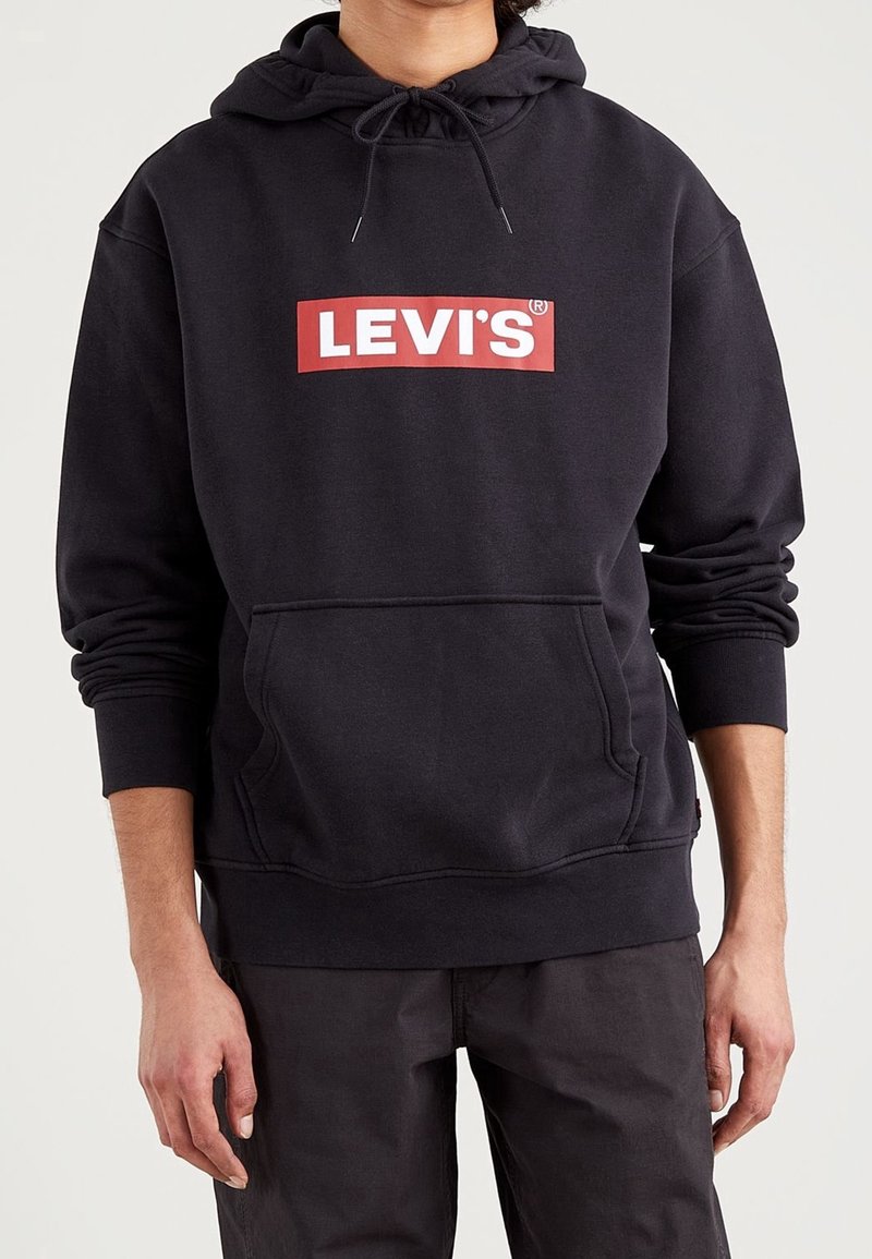 Personne portant un sweat à capuche noir Levi's avec un logo rouge et blanc sur la poitrine et un pantalon foncé assorti, debout les bras détendus.