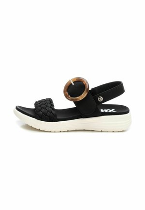 Sandal plana de tejido negro con un acento de hebilla redonda marrón, correa ajustable y suela de goma blanca para tracción y comodidad.