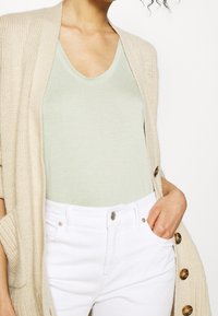 Beige pletený kardigan s bočními kapsami, světle zelené tričko s výstřihem do V, bílé džíny z denimu s detailním zapínáním na knoflík. Viditelná ruka v kapse.