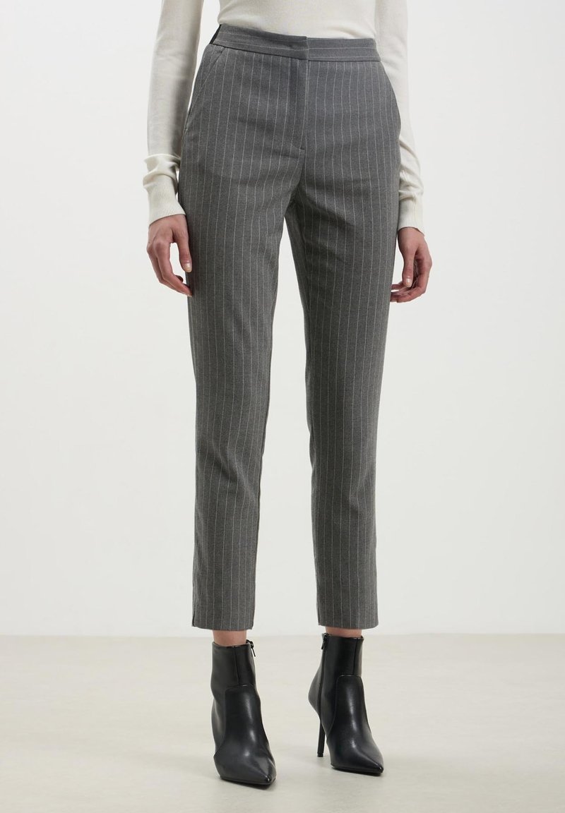 Femme portant un pantalon slim gris à fines rayures, un haut crème à manches longues et des bottines noires à talons hauts, debout sur un sol uni.
