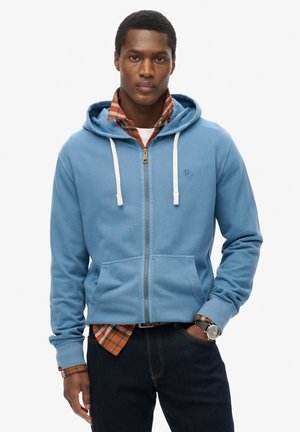 Haut à zip bleu avec des cordons blancs, poches avant et texture douce, porté sur une chemise à carreaux et associé à un jean foncé.