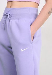 Šviesiai purpurinės sportinės kelnės su atlenkiamu juosmeniu, šoninėmis kišenėmis ir baltu Nike logotipu apatinėje kairėje kojoje. Lygus audinio paviršius.