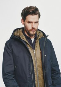 Granatowa kurtka z kapturem, z pikowaną oliwkową warstwą wewnętrzną. Posiada zamek błyskawiczny oraz przednie kieszenie z widocznymi detalami sprzętowymi.