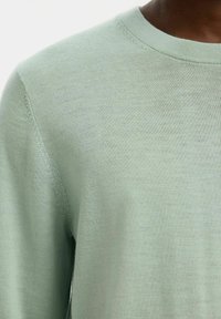 Pull col rond vert clair en tissu doux et texturé. Le gros plan révèle des motifs subtils de tricot et des détails de couture soignés.