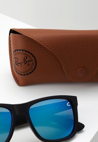Ochelari de soare Ray-Ban cu un cadru negru mat, lentile cu nuanță albastră și un toc din piele maro, care are un logo circular și închidere cu buton.
