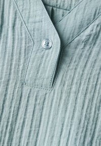 Tessuto strutturato azzurro chiaro con pieghe verticali, caratterizzato da un design a scollo a V e una chiusura con bottone argentato sul davanti.