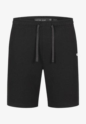 Homme debout face à l'avant portant un t-shirt blanc uni, un short noir et des sandales beige sur un fond gris clair.