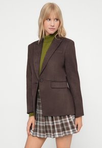 Bruine blazer met een gladde textuur, enkele knop en twee zakken aan de voorkant; gelaagd over een groene coltrui en een geruite rok.
