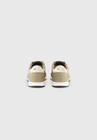 Beige treningssko med teksturert overdel, hvit mellomsåle og svart yttersåle. Funksjoner inkluderer en polstret krage og minimal merkevare.