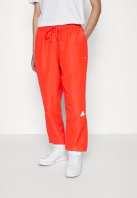 Person som bär ljusröda dragbandbyxor och vita sneakers, står med en hand i fickan mot en enkel vit bakgrund.