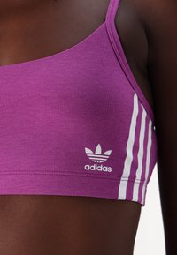 Violet sportinė liemenėlė, pagaminta iš lygaus audinio, su baltais trimis juostelėmis ir adidas logotipu kairėje pusėje po krūtine.