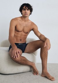 Schwarze Boxerbriefs mit einem weißen Logo haben eine enge Passform. Das Modell hat zerzaustes, lockiges Haar und eine durchtrainierte Figur und sitzt auf einem strukturierten beigen Stuhl.