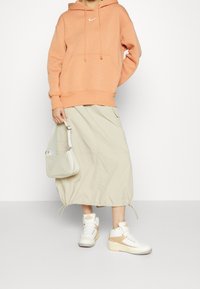 Orange hoodie med en framficka och logotyp, beige elastisk midjeskirt, vita och bruna högskaftade sneakers, samt en liten ljus väska.