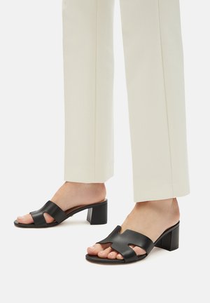 Füße in schwarzen, offenen Sandalen mit Blockabsatz, kombiniert mit weißen, geraden Hosen vor einem schlichten Hintergrund.