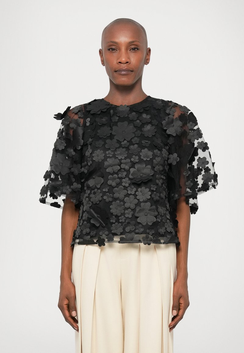Sort blomster-bluse med gennemsigtigt stof og 3D-applikationer, med korte flæseærmer og rund halsudskæring, stylet med lyse bukser.