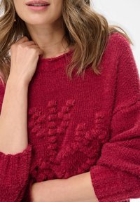 Czerwony sweter o luźnym kroju z szerokim dekoltem i wyraźnymi wzorami dzianiny; wykonany z miękkiego, puszystego materiału.