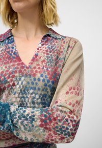Blouse en V légère en tissu imprimé avec des couleurs rose, bleu et neutres ; dotée d'un col et de manches longues.