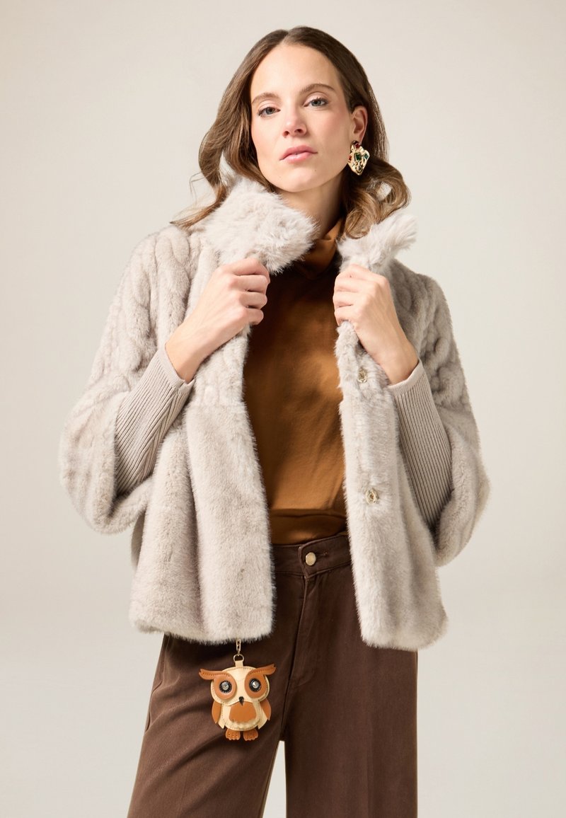 Giacca in pelliccia sintetica grigio chiaro con ampio colletto, motivo strutturato e chiusura con bottoni. Pantaloni marroni a vita alta e portachiavi con gufo.