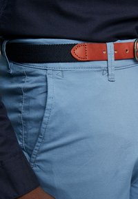 Světle modré kalhoty s hladkou texturou, doplněné černým tkaným pásem a hnědým koženým detailem. U kapes jsou viditelné švy.