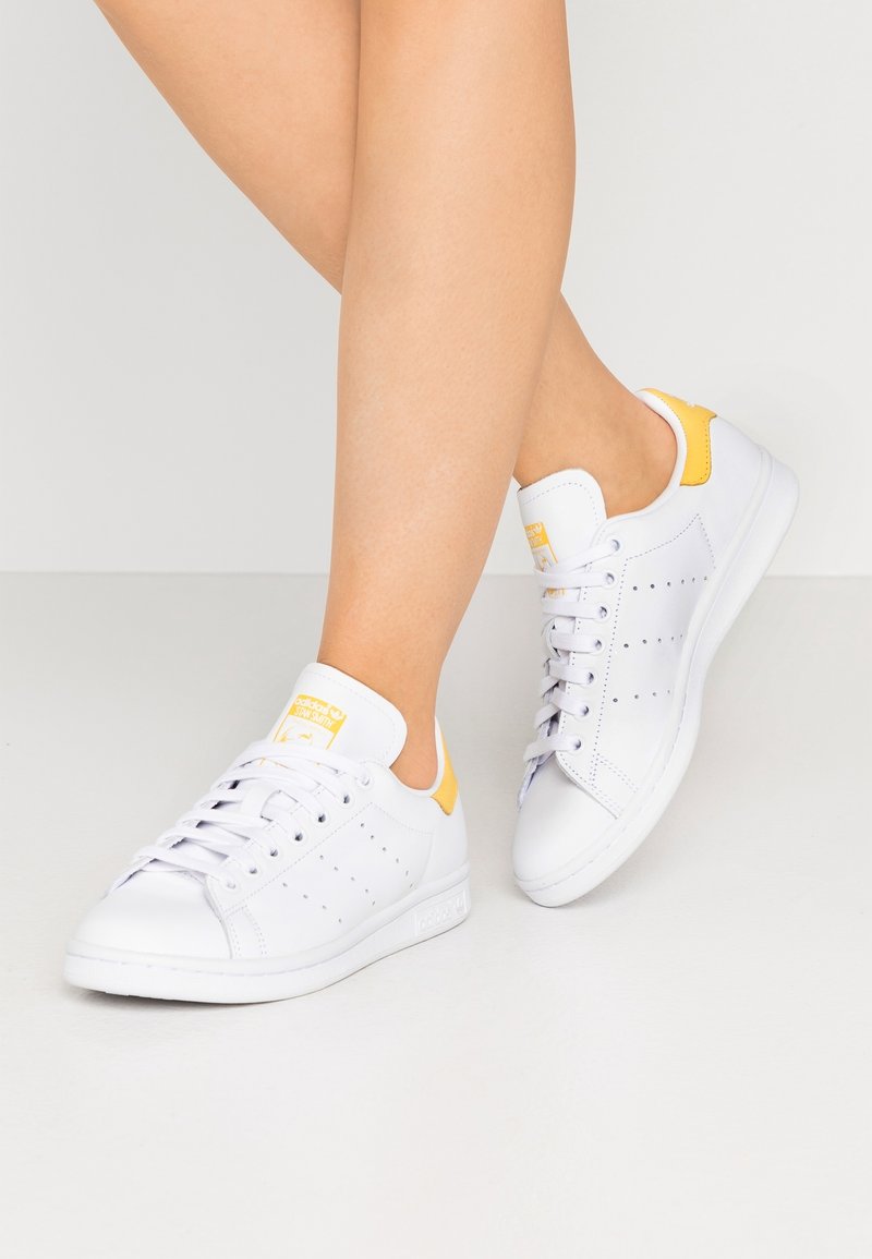 Baskets en cuir blanc avec des accents de talon jaunes, design perforé, semelles en caoutchouc blanc plates et système de laçage standard.