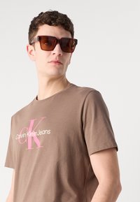 Pruun puuvillane t-särk, millel on roosa "Calvin Klein Jeans" logo. Mudel kannab suuri tumepruune ruudukujulisi prille, mis rõhutavad paksu käte ja kaela.