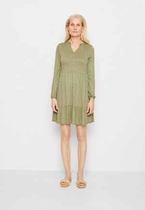 Zelené kvetinové šaty s dlhými rukávmi, výstrihom do V a volánovým lemom; ľahký materiál s rozptýlenými ružovými a modrými kvetmi. Nosí sa s plochými vrkôčkovými sandálmi.