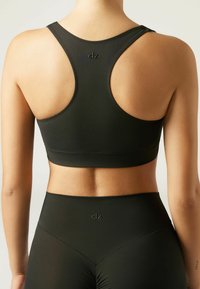 Schwarzer Sport-BH mit Racerback-Design, glatter Textur und dezentem Branding. Das Material hat eine matte Oberfläche und passt gut zu hochgeschnittenen Leggings.
