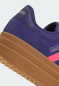 Gros plan d'une basket Adidas violette avec semelle en gomme, bande rose, surface texturée et logo Adidas sur le talon et la semelle.