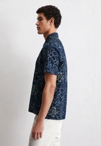 Korte mouwen overhemd van marineblauwe stof met bloemmotieven in lichtgroen, zachte textuur met een kraag, weergegeven op een model vanuit de zijkant.