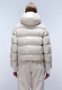Giacca a vento color crema con colletto alto, design trapuntato e finitura lucida. Indossata con pantaloni chiari, vista da dietro.