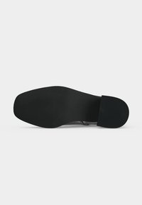 Semelle de chaussure en cuir noir avec un bout carré et un petit talon bloc reposant sur une surface blanche.