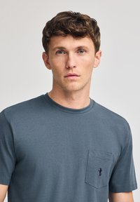 Camiseta de algodón azul oscuro con cuello redondo, mangas cortas y un bolsillo en el pecho izquierdo que presenta un logo bordado de una figura.