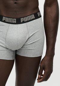 Torso masculin et parties supérieures des jambes portant des boxers Puma gris avec une ceinture noire et le logo Puma, bras droit détendu sur le côté.