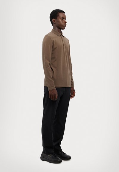 Polo cu mânecă lungă, de culoare maro, dintr-un material neted, asortat cu pantaloni negri și adidași negri, având un design minimalist.