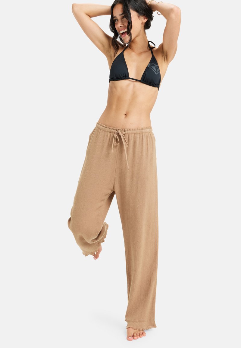 Beige, könnyű, bőszárú nadrág, húzózsinórral a deréknál és rojtos szegélyekkel. Fekete bikini toppal párosítva. Simaszerű textúra.